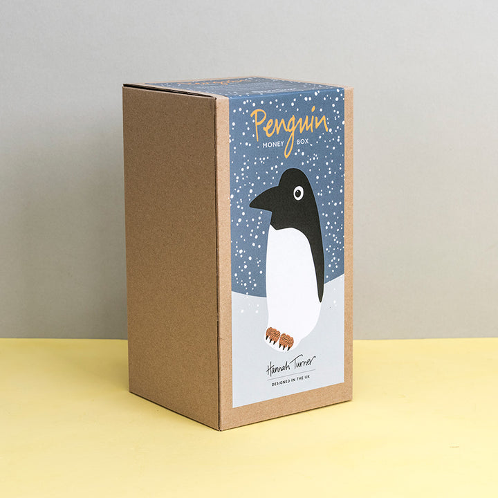 Brown kraft gift box with penguin design on a grey snowy background printed with text 'Hannah Turner designed in the UK'. Packaging for Hannah Turner's Ceramic Penguin Money Box.