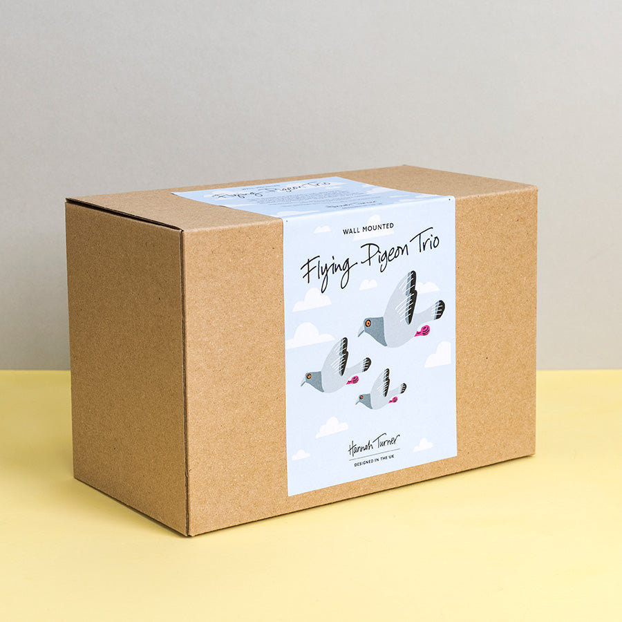 Brown kraft box with flying pigeon design on a light sky blue background with clouds and printed with text 'Hannah Turner designed in the UK'. Packaging for Hannah Turner's Flying Pigeon Trio Ceramic Wall Art.