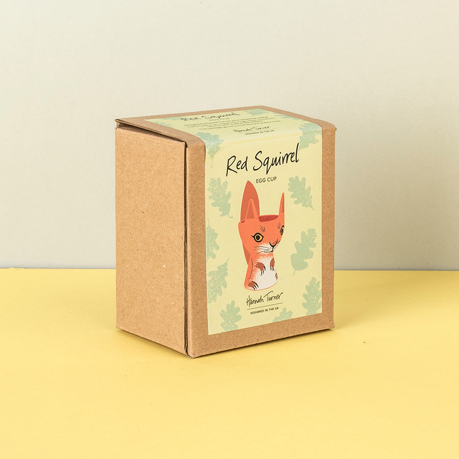 A square kraft paper box for a 'Red Squirrel Egg Cup' by 'Hannah Turner', featuring an illustration of a red squirrel on a cream background with light green leaf motifs, set against a yellow surface and a light gray wall.