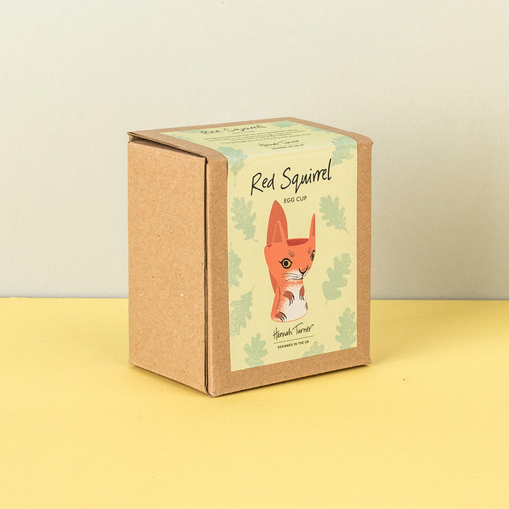 A square kraft paper box for a 'Red Squirrel Egg Cup' by 'Hannah Turner', featuring an illustration of a red squirrel on a cream background with light green leaf motifs, set against a yellow surface and a light gray wall.