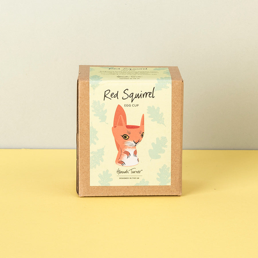 A square kraft paper box for a 'Red Squirrel Egg Cup' by 'Hannah Turner', featuring an illustration of a red squirrel on a cream background with light green leaf motifs, set against a yellow surface and a light gray wall.
