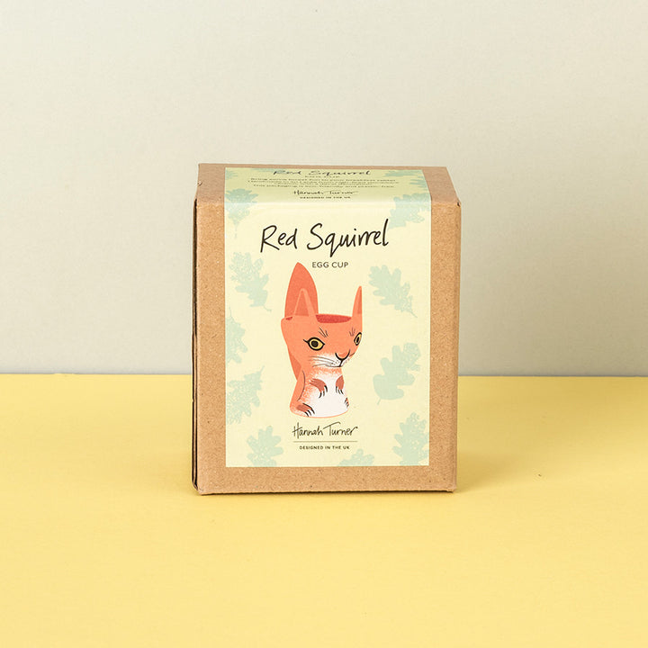 A square kraft paper box for a 'Red Squirrel Egg Cup' by 'Hannah Turner', featuring an illustration of a red squirrel on a cream background with light green leaf motifs, set against a yellow surface and a light gray wall.