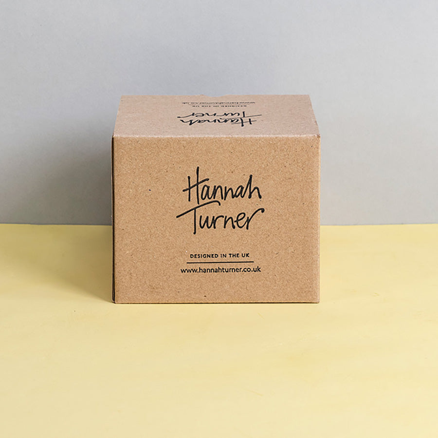 A square kraft paper box standing upright on a pale yellow surface against a light grey background. The front of the box is printed with the brand name Hannah Turner in black script and smaller text below reading 'Designed in the UK' and Hannah Turner's website address.