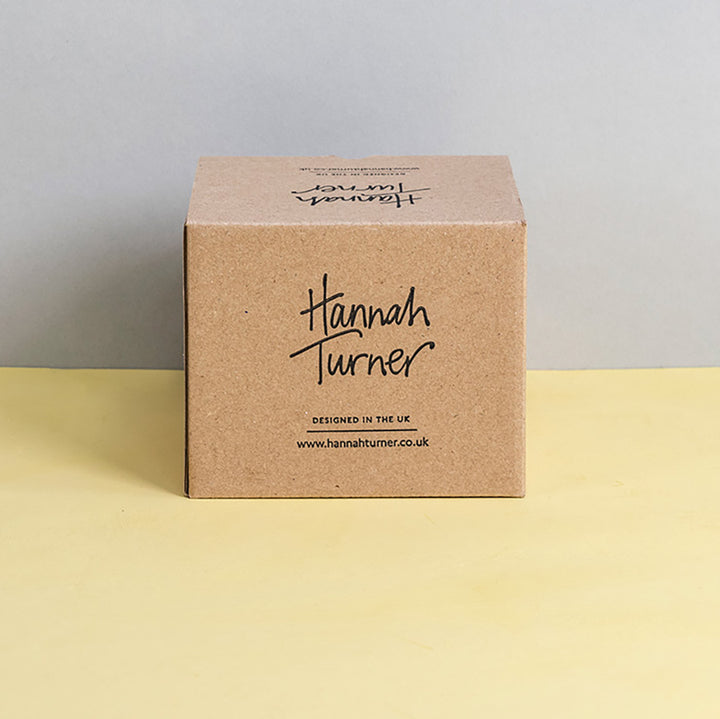 A square kraft paper box standing upright on a pale yellow surface against a light grey background. The front of the box is printed with the brand name Hannah Turner in black script and smaller text below reading 'Designed in the UK' and Hannah Turner's website address.