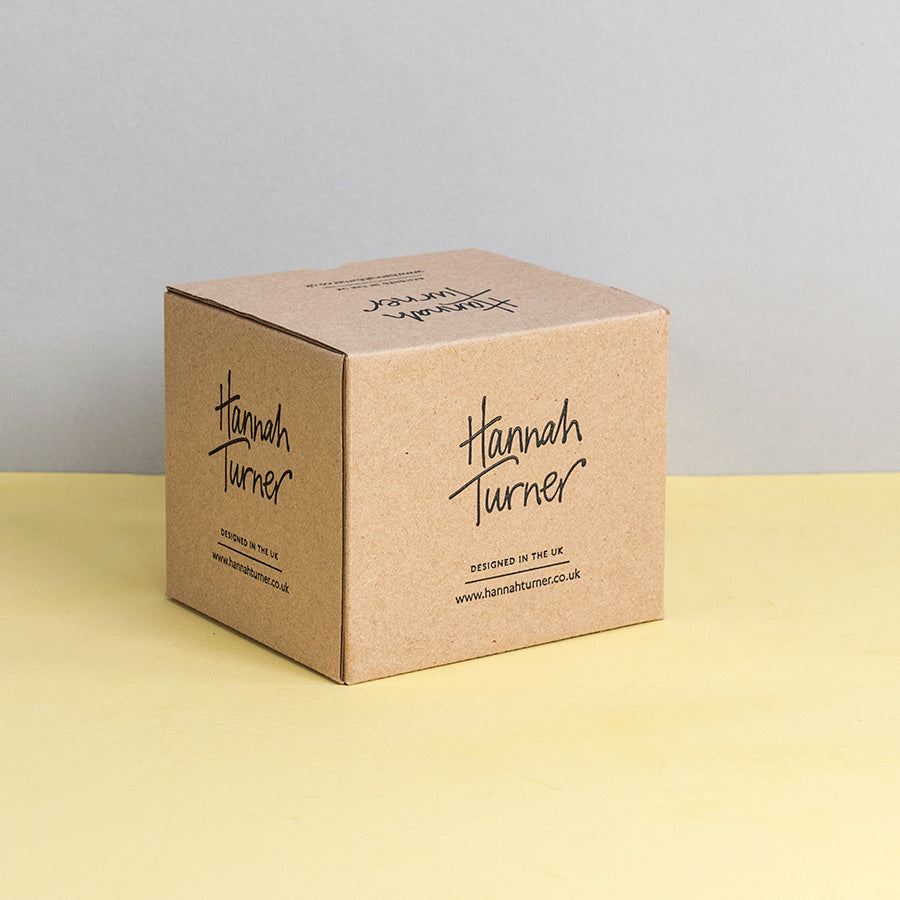 A square kraft paper box standing upright on a pale yellow surface against a light grey background. The front of the box is printed with the brand name Hannah Turner in black script and smaller text below reading 'Designed in the UK' and Hannah Turner's website address.