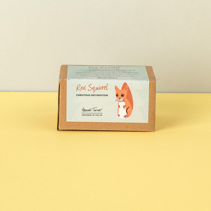 A brown kraft paper box for a 'Red Squirrel Christmas Decoration' by Hannah Turner, featuring a light blue label with an illustration of a red squirrel, set against a split beige and yellow background.