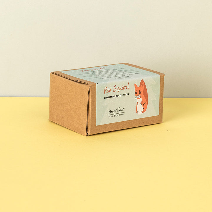 A small brown kraft paper box with a printed label reading 'Red Squirrel Christmas Decoration' and showing an illustration of a red squirrel, photographed on a pale yellow surface against a light beige wall.