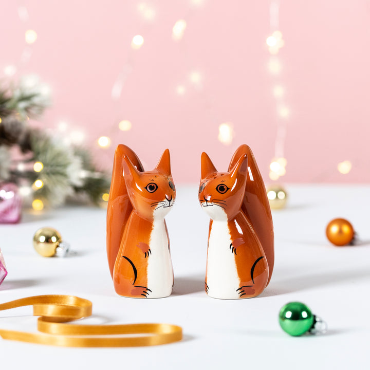 Two small ceramic squirrel salt and pepper shakers in orange and white, standing side by side on a white surface with a soft pink background, holiday lights, ornaments, and a ribbon.