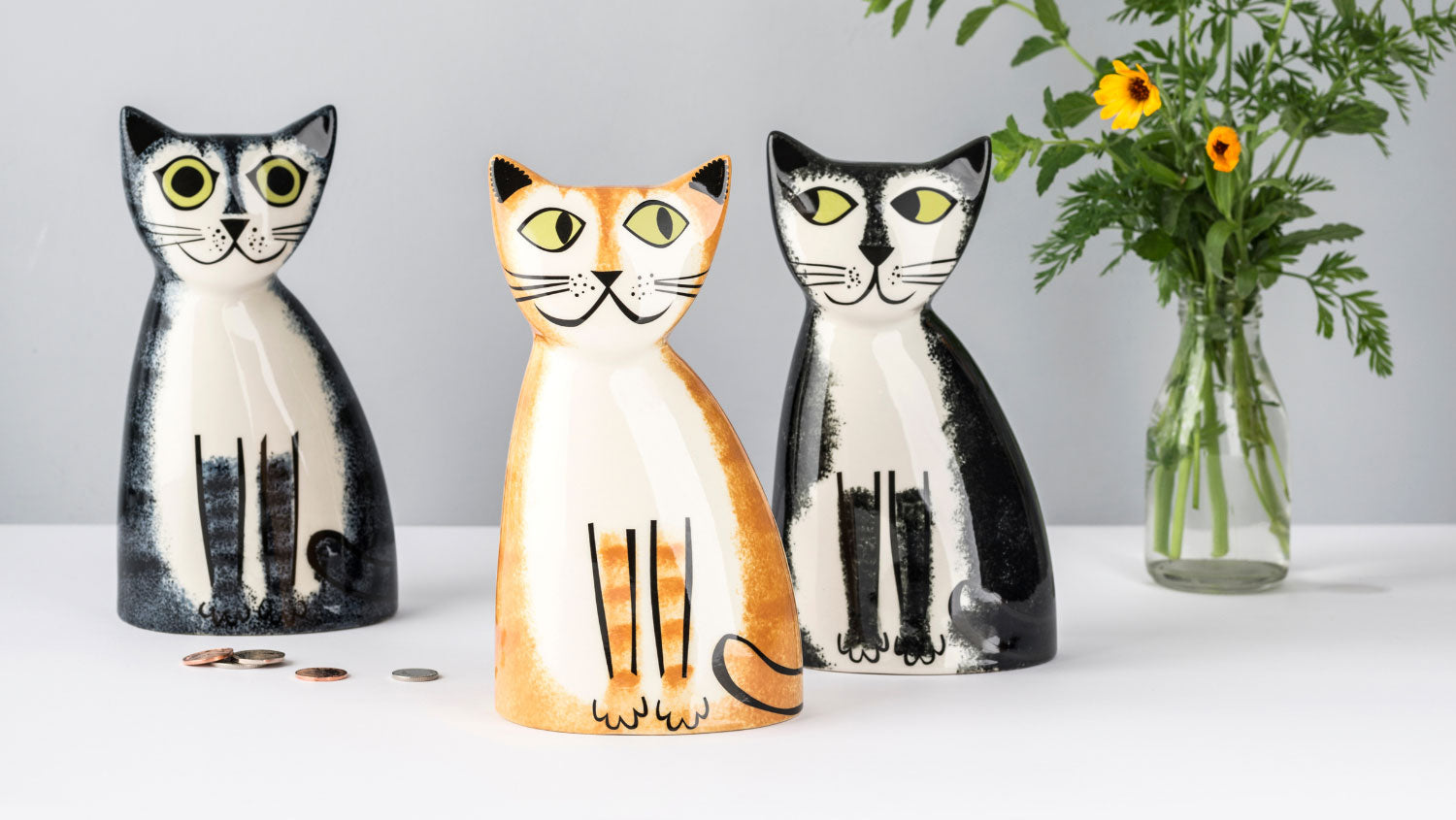 Three ceramic cat-shaped pig banks in black and white, orange and cream, and black and white designs, arranged on a white surface with a glass vase of flowers in the background.