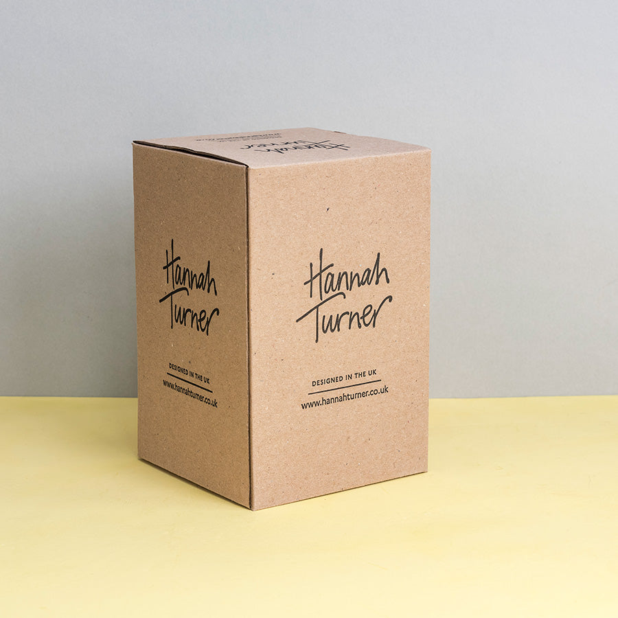 A square kraft paper box standing upright on a pale yellow surface against a light grey background. The front of the box is printed with the brand name Hannah Turner in black script and smaller text below reading 'Designed in the UK' and Hannah Turner's website address.