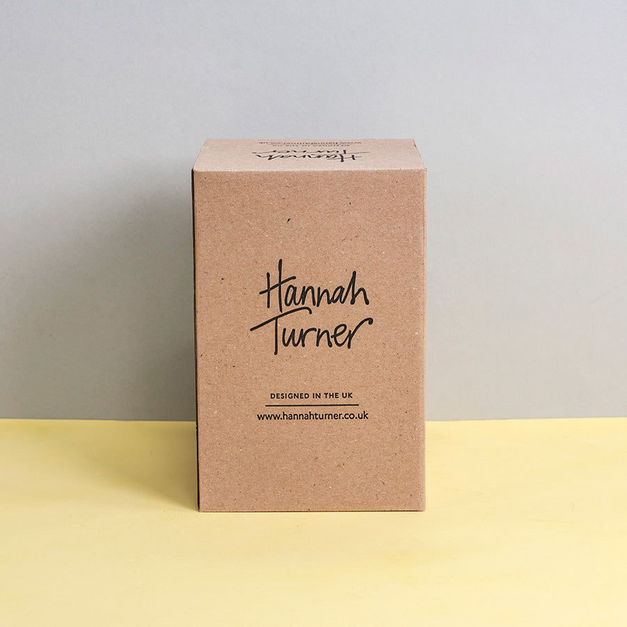 A square kraft paper box standing upright on a pale yellow surface against a light grey background. The front of the box is printed with the brand name Hannah Turner in black script and smaller text below reading 'Designed in the UK' and Hannah Turner's website address.