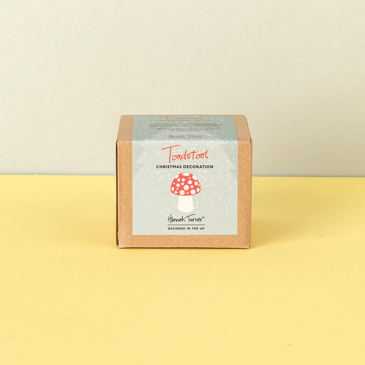 A square kraft paper box with a snowy illustrated label reading 'Toadstool Christmas Decoration ' and 'Handmade in the uk' photographed on a pale yellow surface against a light beige wall.