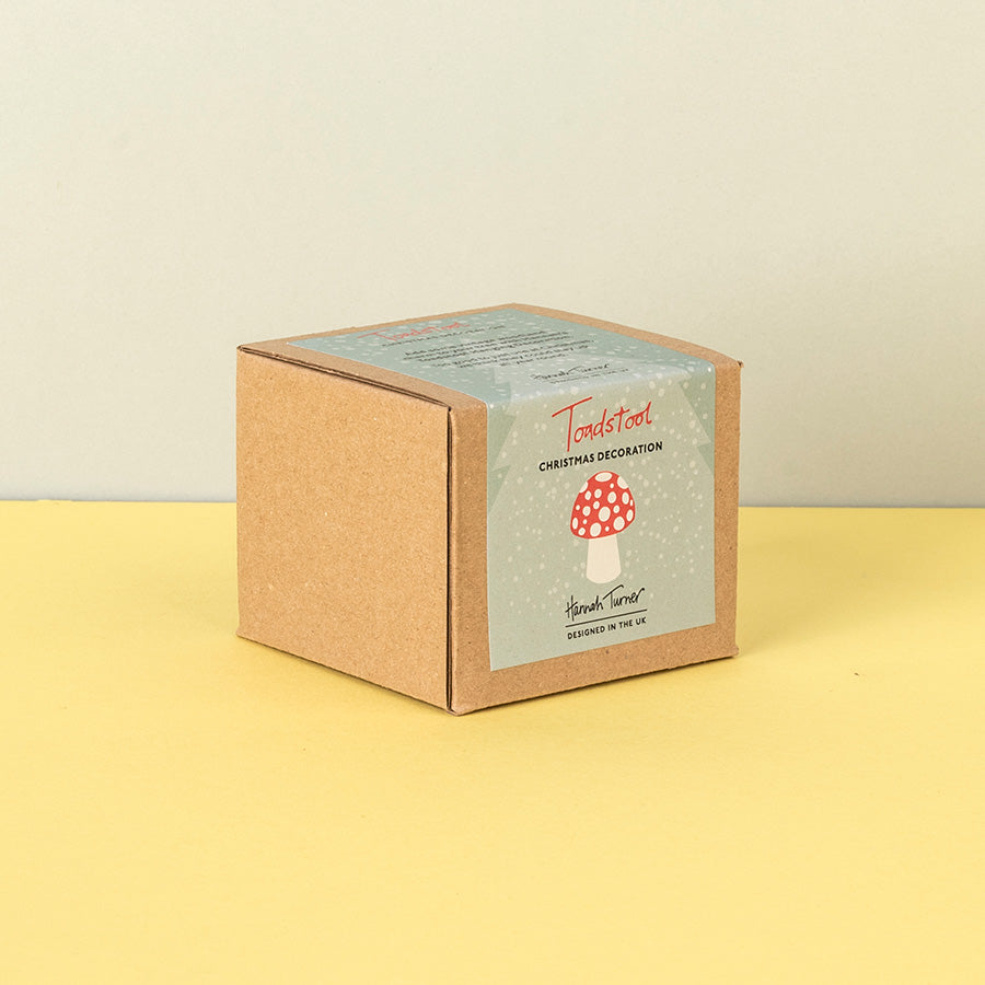 A square kraft paper box with a snowy illustrated label reading 'Toadstool Christmas Decoration ' and 'Handmade in the uk' photographed on a pale yellow surface against a light beige wall.