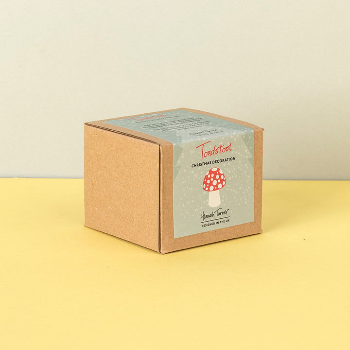 A square kraft paper box with a snowy illustrated label reading 'Toadstool Christmas Decoration ' and 'Handmade in the uk' photographed on a pale yellow surface against a light beige wall.