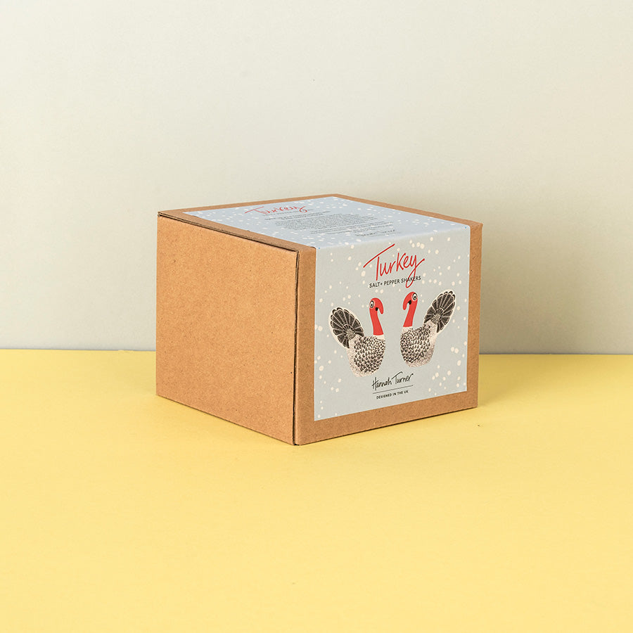 A square kraft paper box with a white front label featuring two illustrated turkeys and the text 'Turkey' and Salt & Pepper Shakers standing on a yellow surface against a light beige wall.
