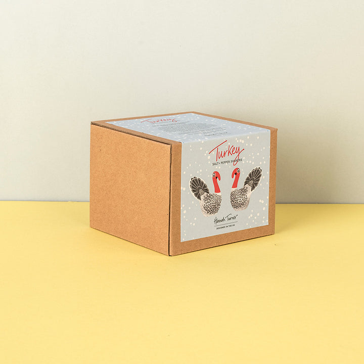 A square kraft paper box with a white front label featuring two illustrated turkeys and the text 'Turkey' and Salt & Pepper Shakers standing on a yellow surface against a light beige wall.