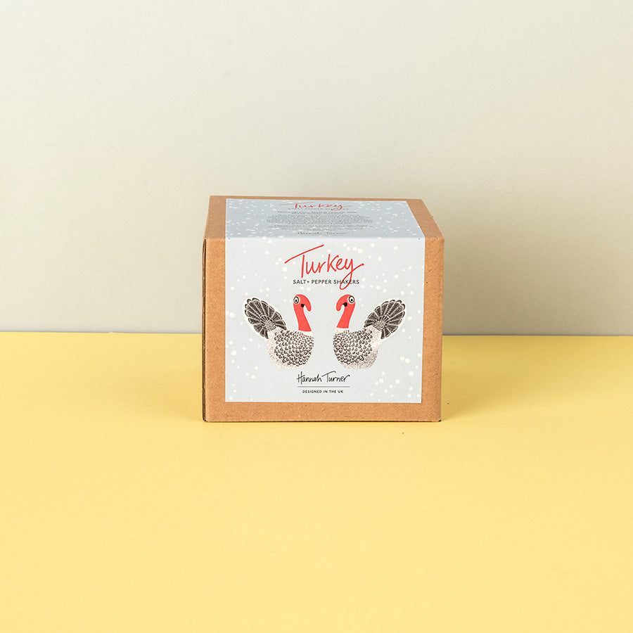 A square kraft paper box with a white front label featuring two illustrated turkeys and the text 'Turkey' and Salt & Pepper Shakers standing on a yellow surface against a light beige wall.