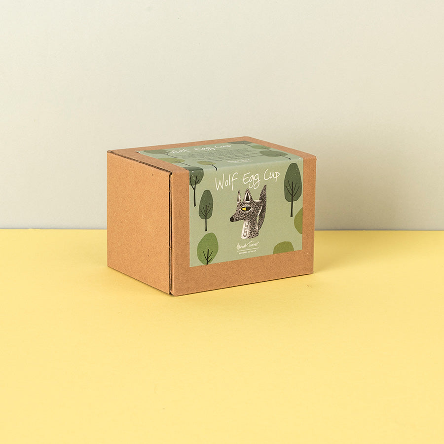A square kraft paper box with a printed label reading 'WOLF Egg Cup' and showing a wolf illustration on a green background, placed on a yellow surface against a light beige wall.