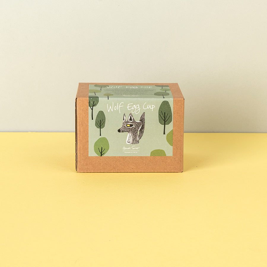 A square kraft paper box with a front label reading 'Wolf Egg Cup' and featuring a wolf illustration, standing on a yellow surface against a light beige wall.