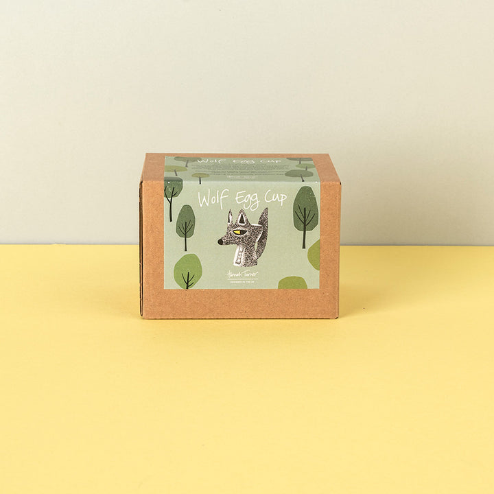 A square kraft paper box with a front label reading 'Wolf Egg Cup' and featuring a wolf illustration, standing on a yellow surface against a light beige wall.