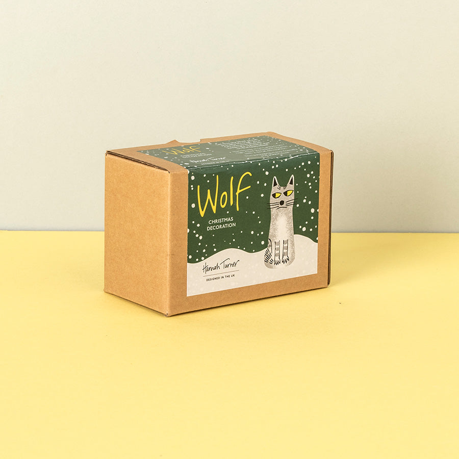 A square kraft paper gift box with a green and white illustrated label reading 'Wolf Christmas decoration' and showing a wolf illustration, standing on a yellow surface against a light beige wall.