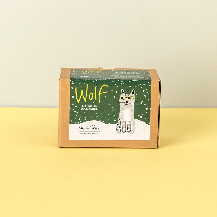 A square kraft paper box for Hannah Turner Wolf Christmas Decoration, featuring a green label with a white wolf illustration and the word 'Wolf' in yellow, set against a yellow and beige background.