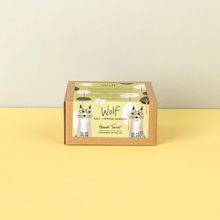 A square cardboard box of Wolf Salt & Pepper Shakers by Hannah Turner, featuring a whimsical illustration of two cats dressed as salt and pepper shakers on a light green label, set against a split yellow and beige background.
