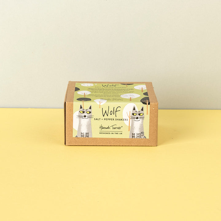 A square cardboard box of Wolf Salt & Pepper Shakers by Hannah Turner, featuring a whimsical illustration of two cats dressed as salt and pepper shakers on a light green label, set against a split yellow and beige background.