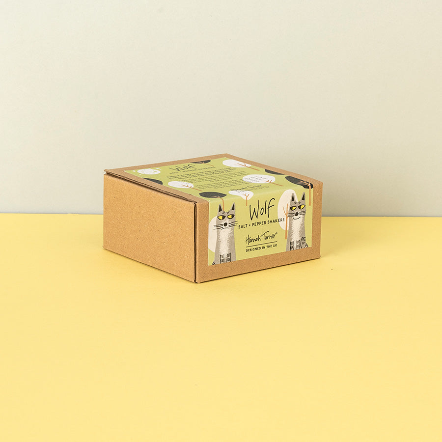 A square kraft cardboard box with a green and white label featuring Wolves and the text 'Wolf Salt - Pepper Shakers', photographed on a yellow surface against a light beige wall.