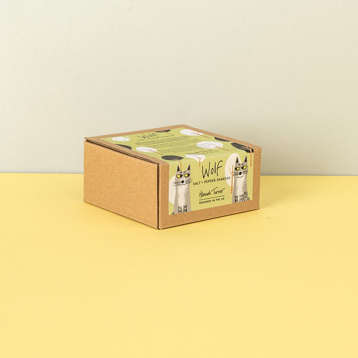 A square kraft cardboard box with a green and white label featuring Wolves and the text 'Wolf Salt - Pepper Shakers', photographed on a yellow surface against a light beige wall.