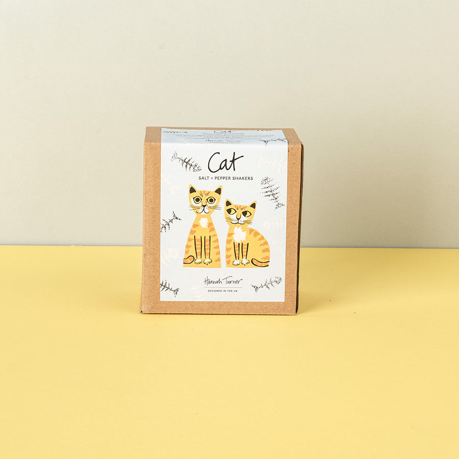 A square kraft paper box for 'Cat Salt + Pepper Shakers' by Handmade Things, featuring an illustration of two orange tabby cats on a white label, set against a split beige and yellow background.