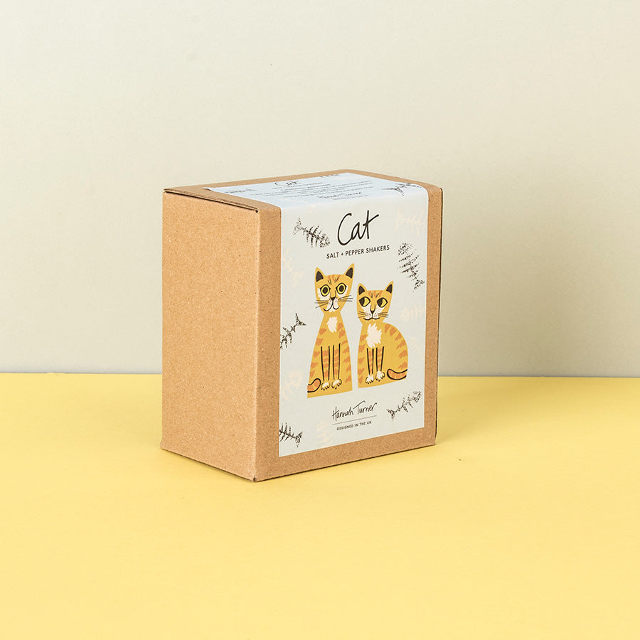 A square kraft paper box with a white label featuring two illustrated orange-and-white cats and the word 'Cat' in script, standing on a yellow surface against a light beige wall.