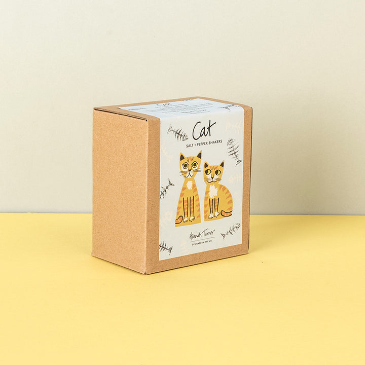 A square kraft paper box with a white label featuring two illustrated orange-and-white cats and the word 'Cat' in script, standing on a yellow surface against a light beige wall.